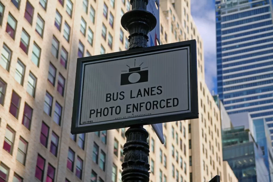 Close-up of "Bus Lanes - Photo Enforced" sign with camera logo on New York City street pole.